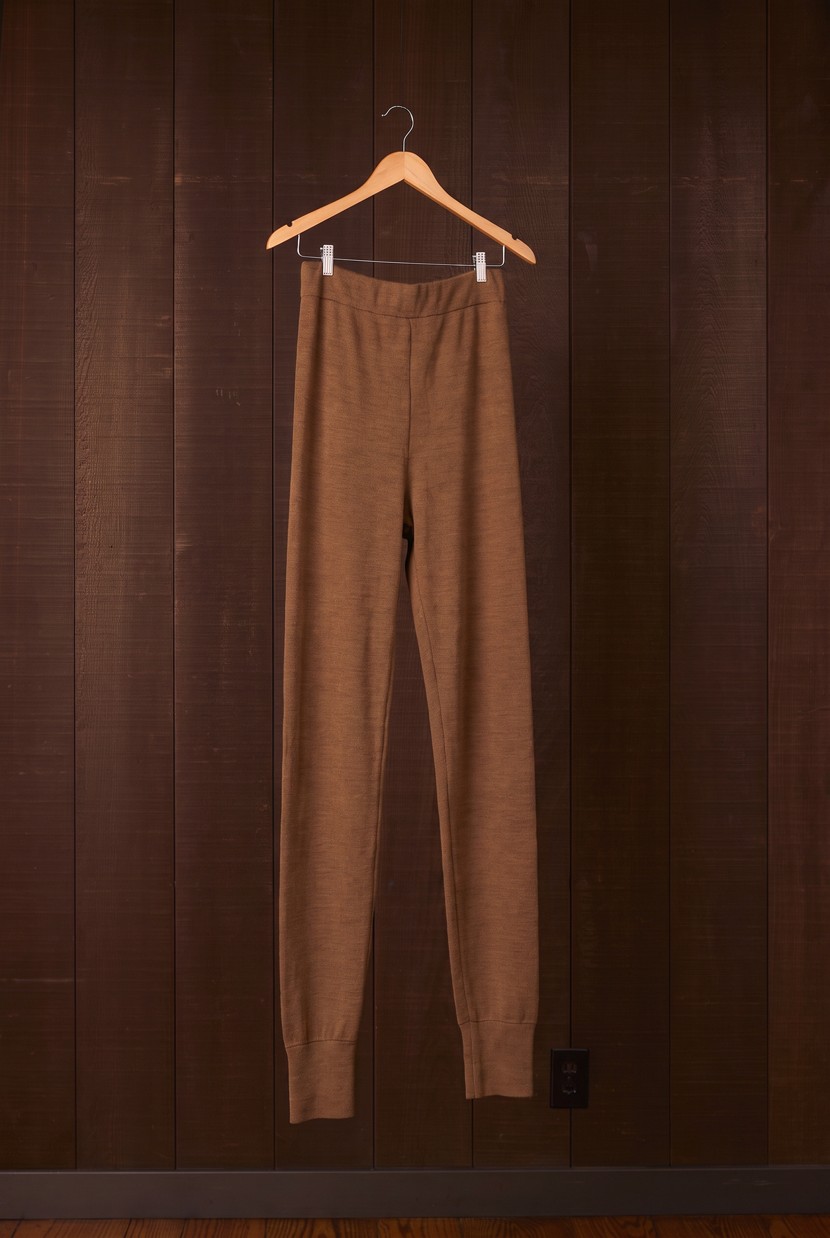 Tall women's walnut brown merino wool leggings on a hanger against a dark wooden wall