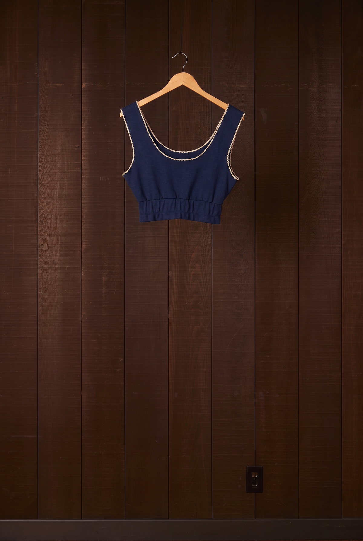 Indigo organic cotton crop tank with cream shell-stitch edging on a hanger against a dark wooden wall