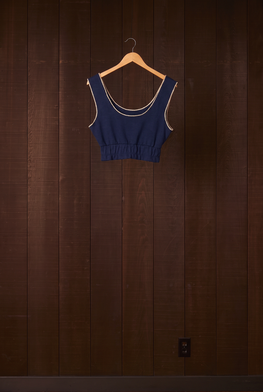 Indigo organic cotton crop tank with cream shell-stitch edging on a hanger against a dark wooden wall