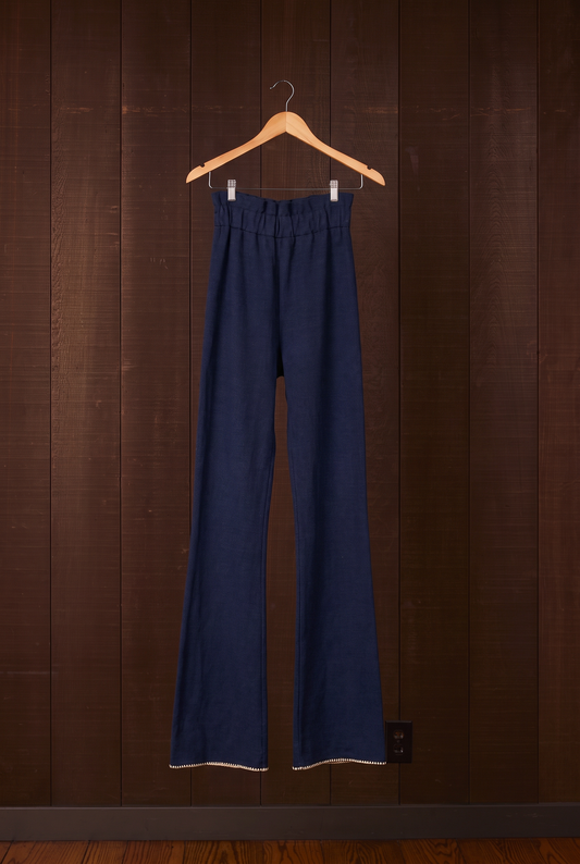 Tall women's indigo organic cotton flare pants with cream shell-stitch hem on a hanger against a dark wooden wall