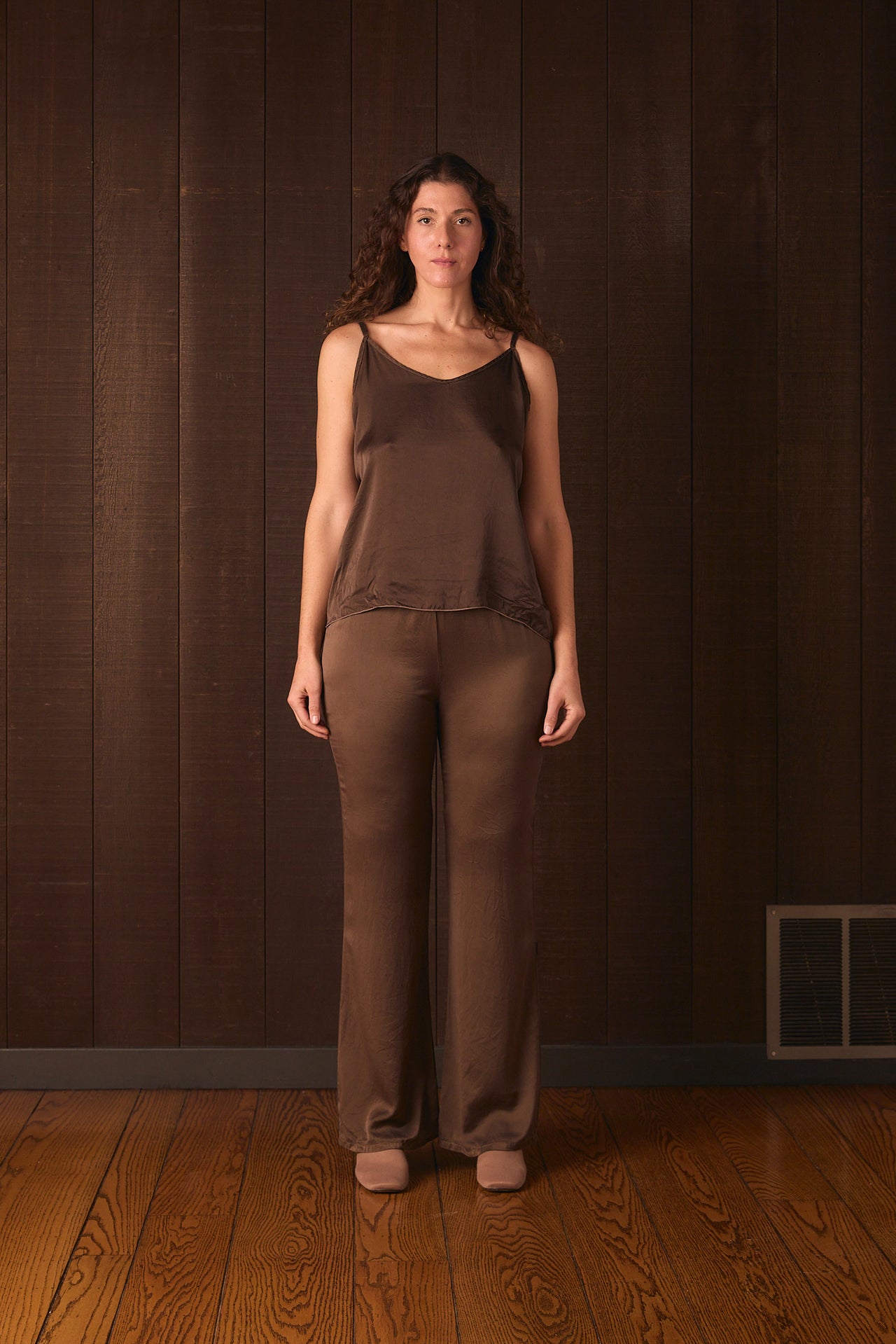 Tall woman wearing a brown sleeveless silk top and pants standing against a dark brown wall.