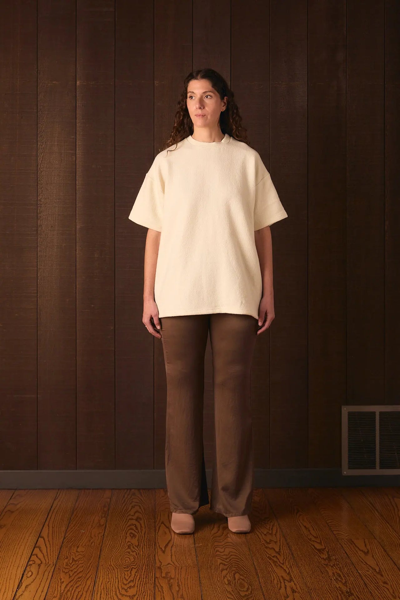 Tall woman wearing a cream sweater and brown pants standing against a wooden paneled wall.
