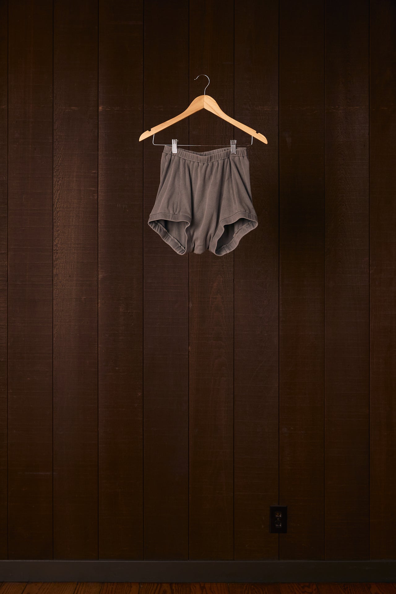 Gray boxer briefs hanging on a wooden hanger against a dark wooden wall.