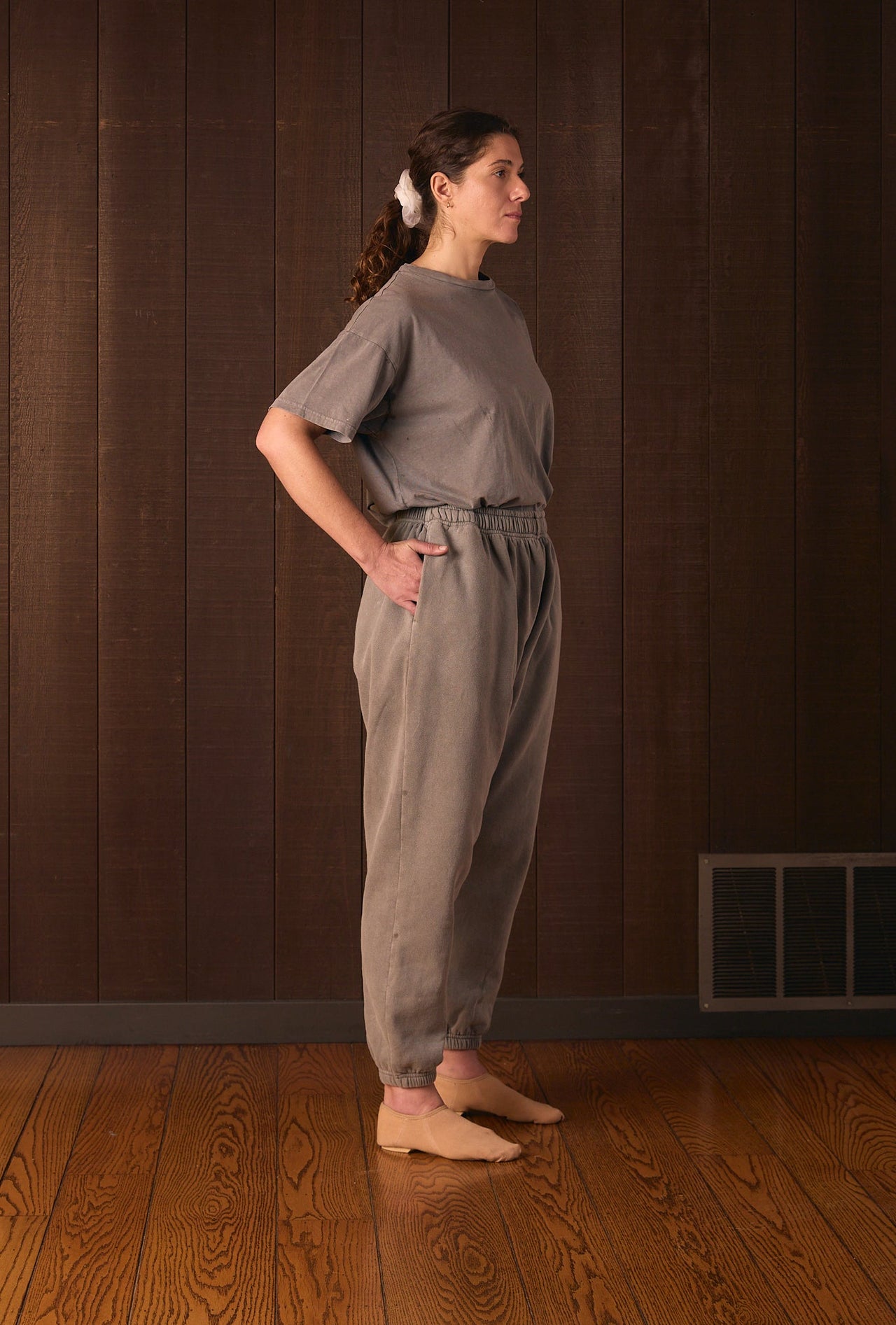 Tall woman wearing a gray jumpsuit standing against a brown wall.