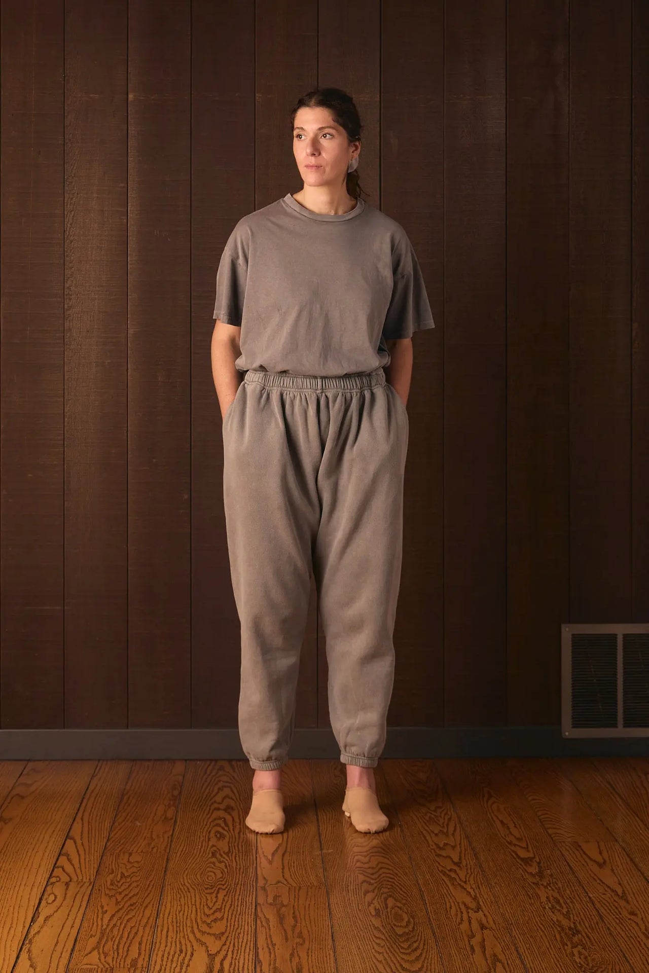 Tall woman wearing a gray outfit standing against a brown wall.