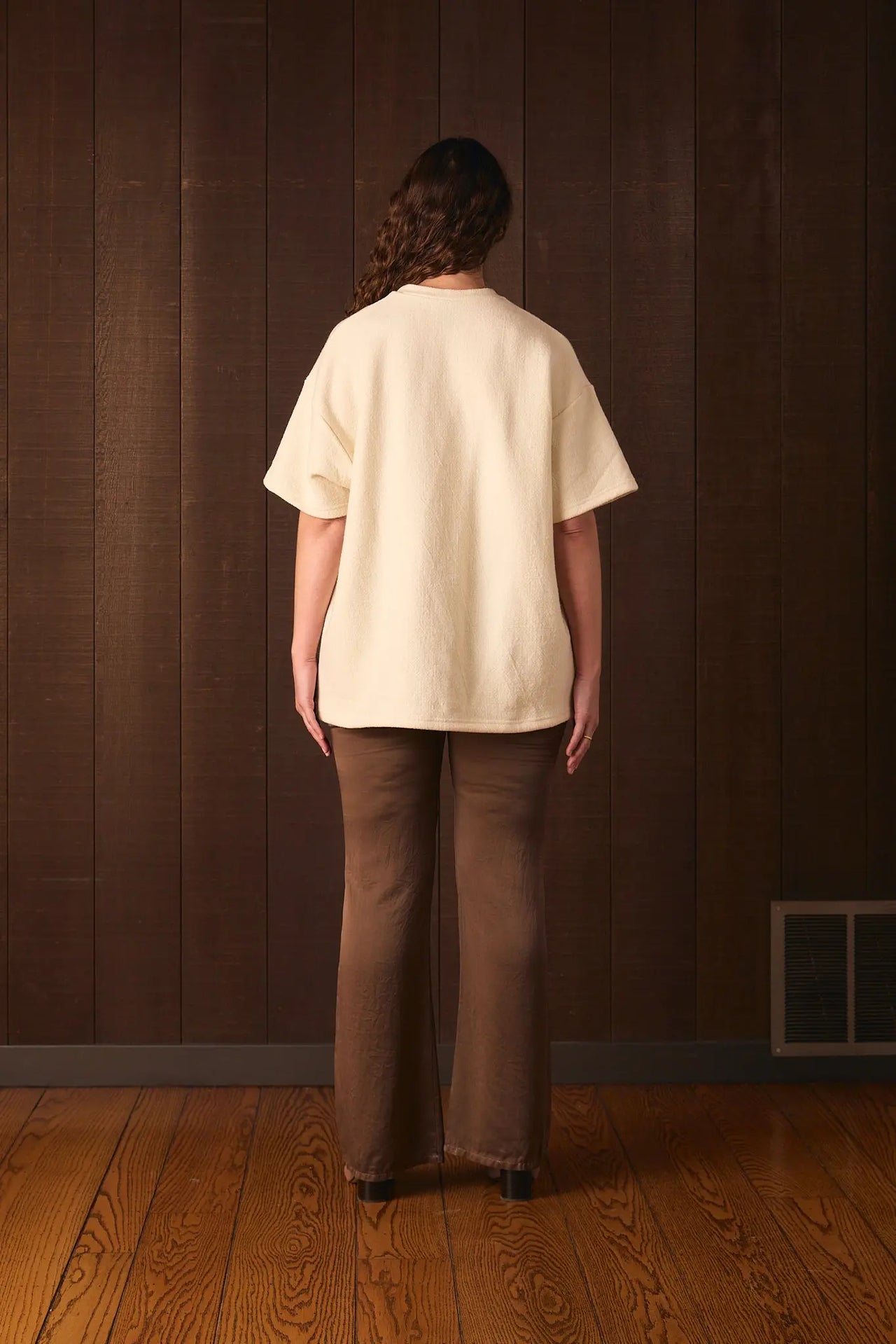 Tall woman wearing a cream-colored shirt and brown pants standing against a wooden paneled wall.