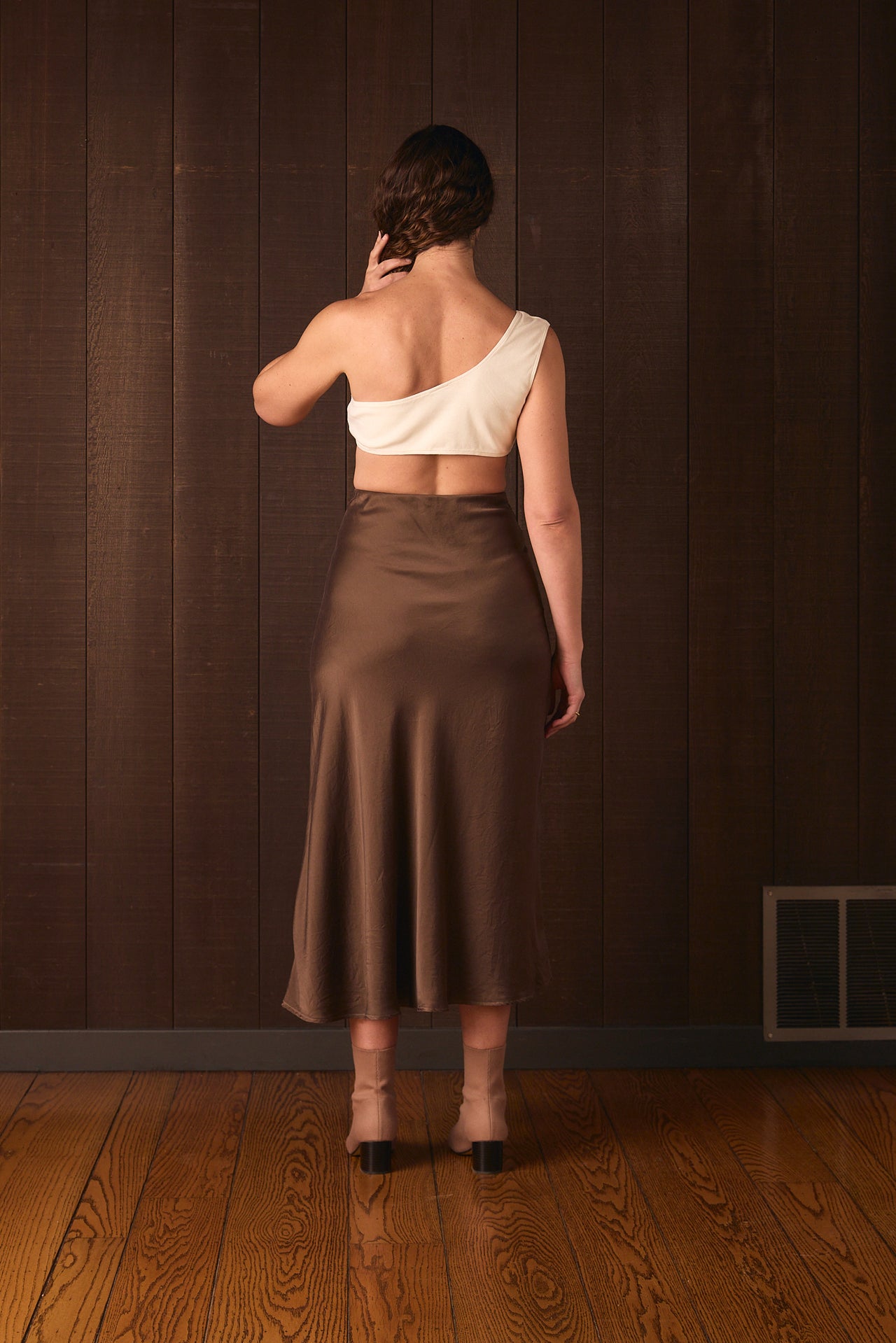 Tall woman wearing a white top and brown skirt against a wooden paneled wall.