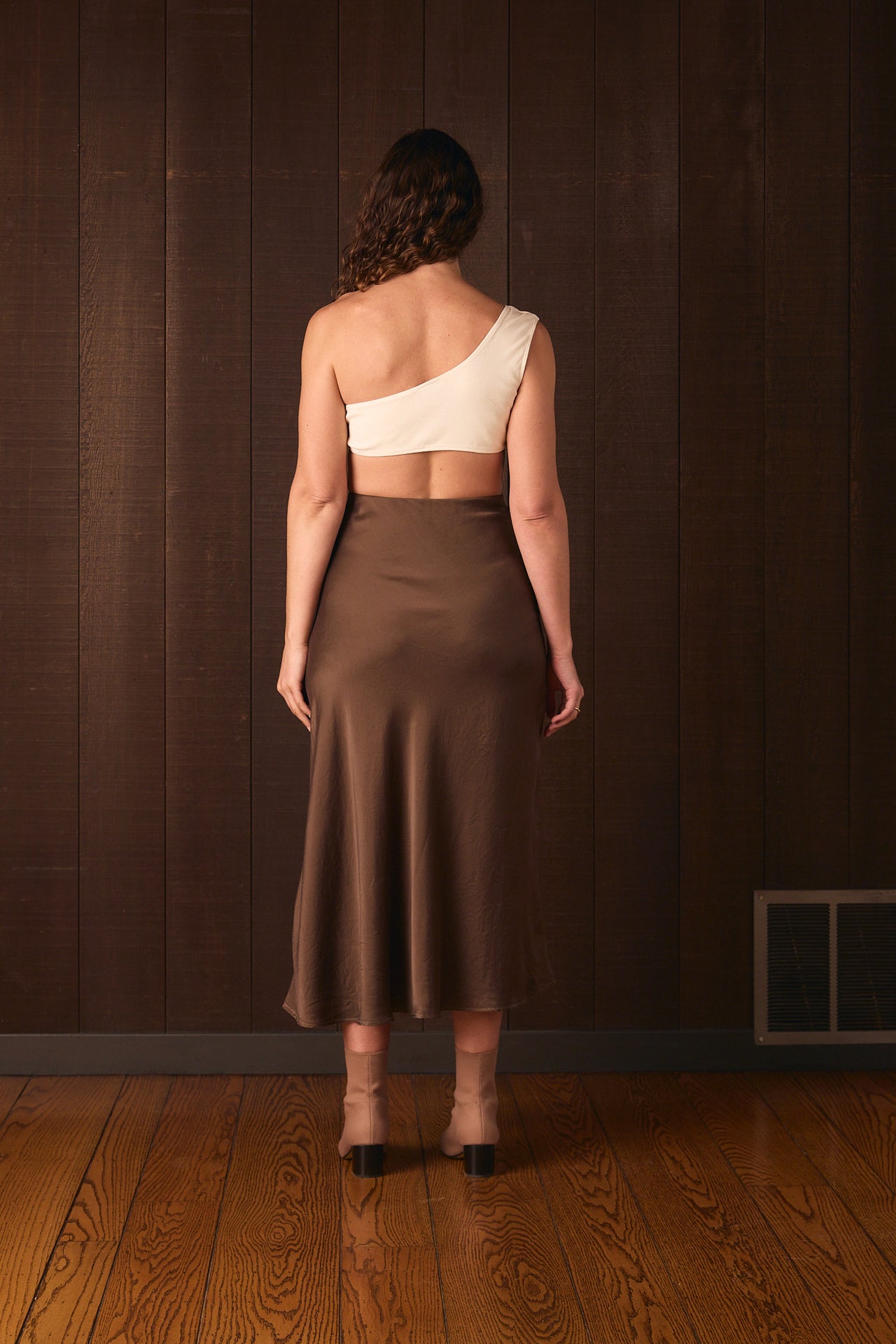 Tall woman wearing a white top and brown skirt standing against a dark wall.
