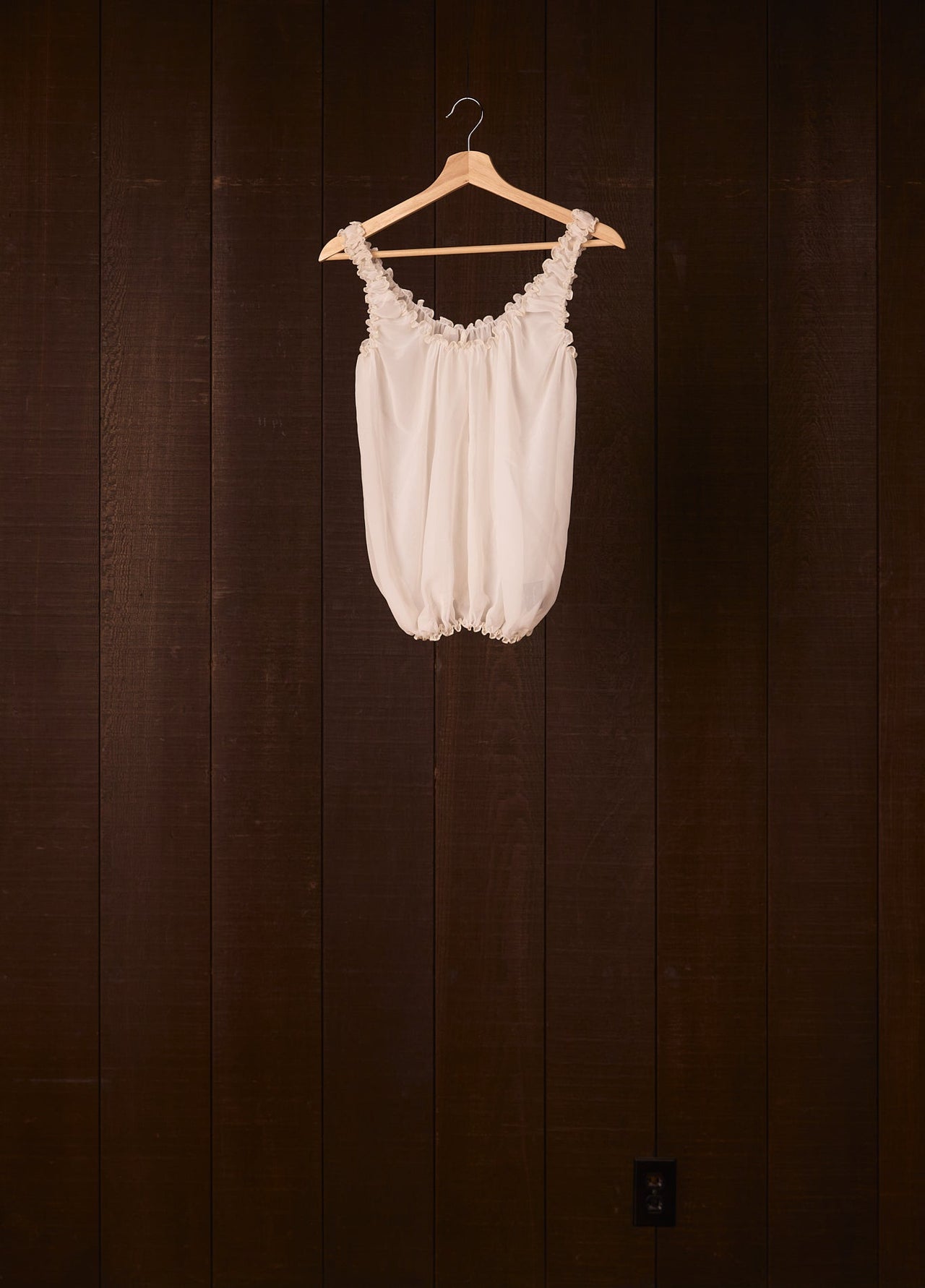 White silk top on a wooden hanger against a dark wooden wall.