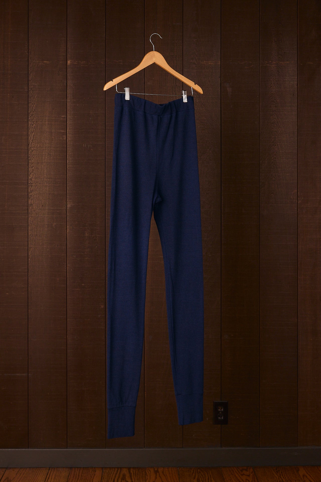 Herderin indigo blue pants hanging on a wooden hanger against a brown striped wall.