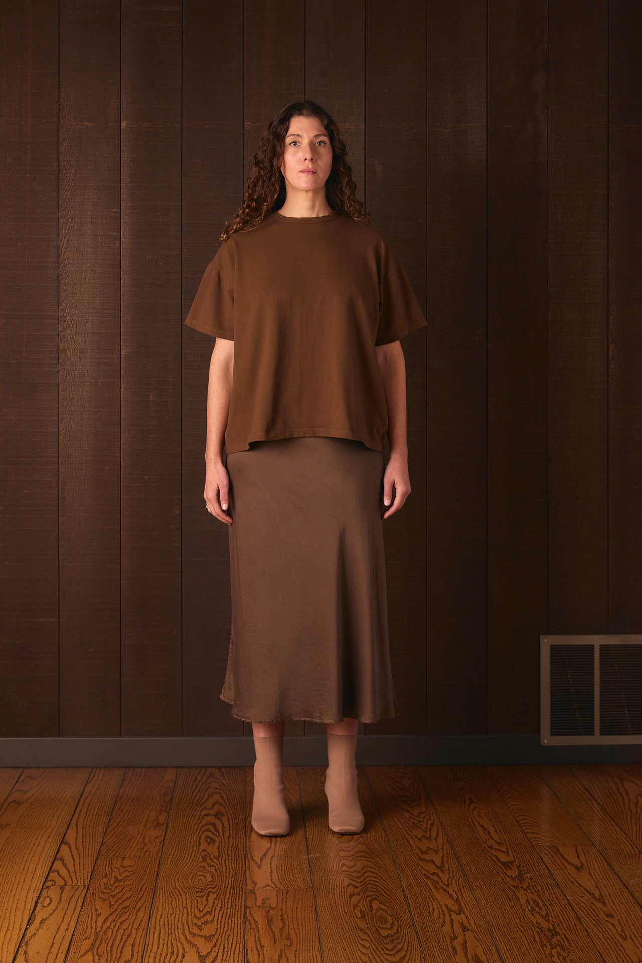 Tall woman wearing a brown outfit standing against a dark wooden wall.