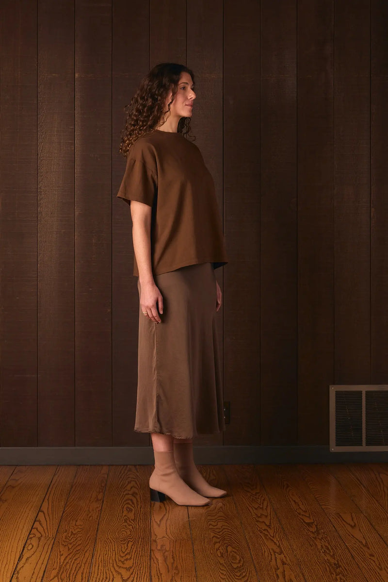 Tall woman wearing a brown outfit standing against a wooden paneled wall.
