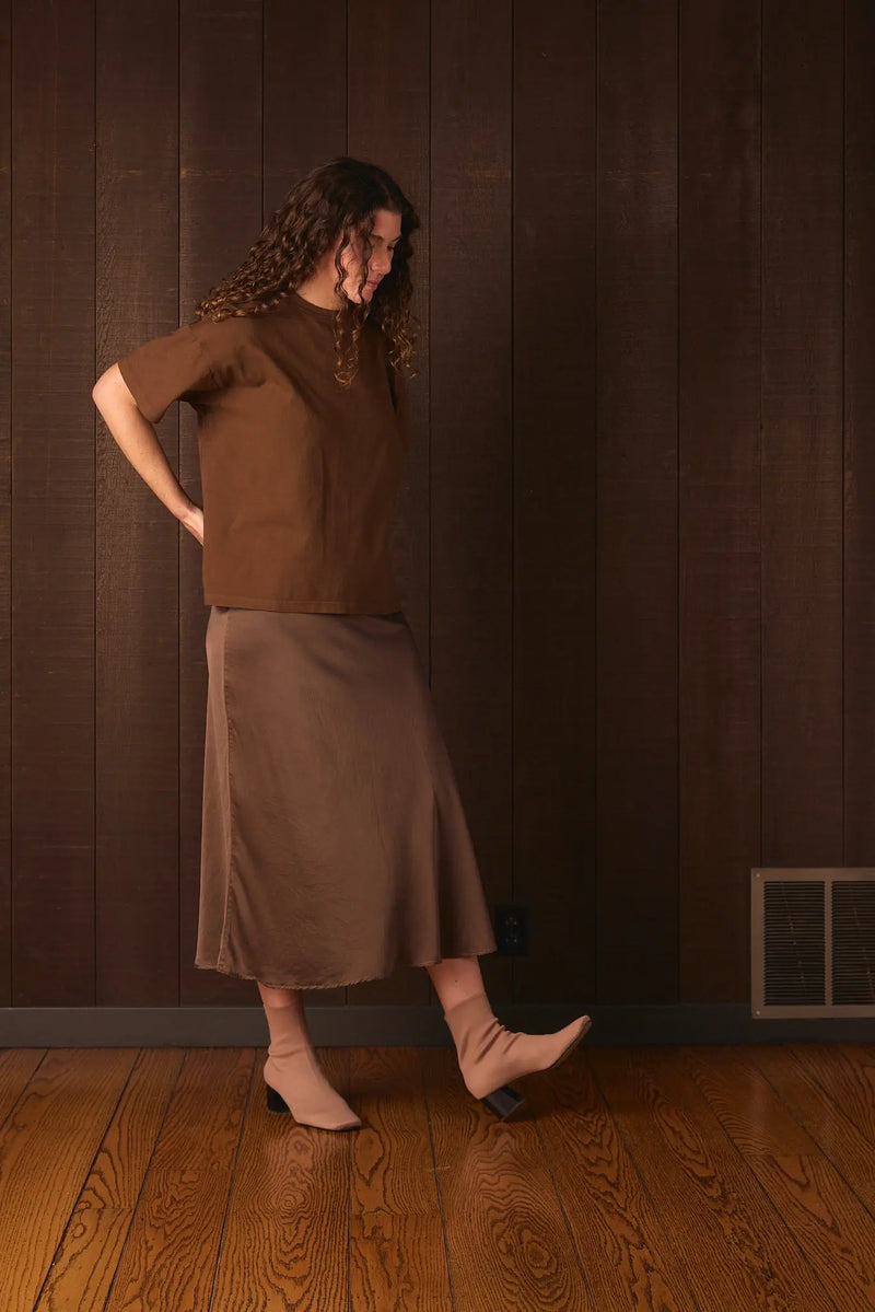 Tall woman wearing a brown outfit standing against a wooden paneled wall.