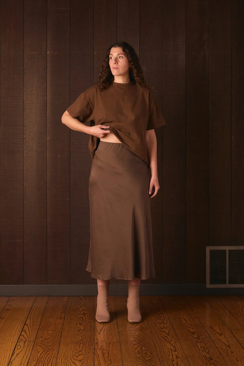Tall woman wearing a brown outfit standing against a dark wooden wall.