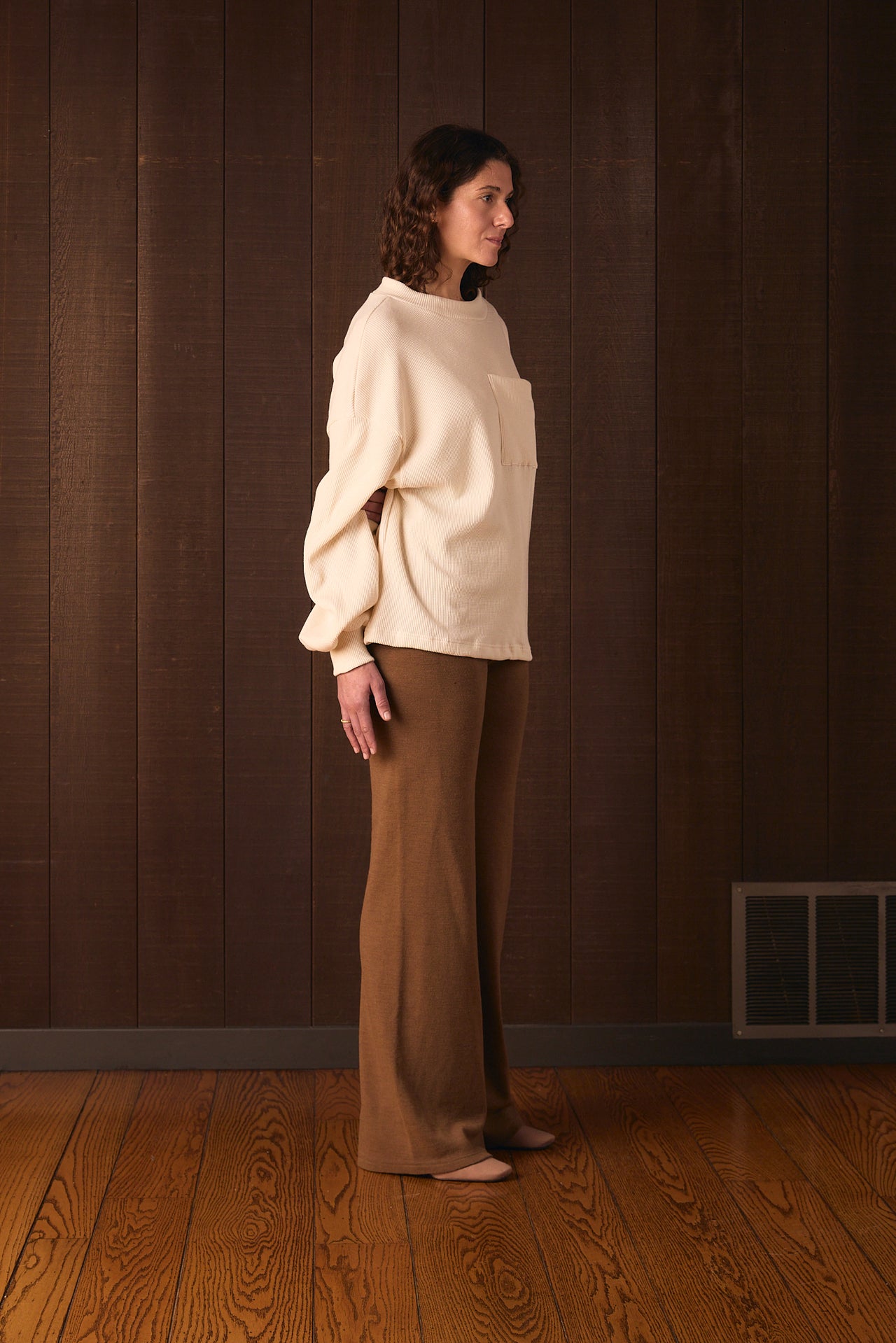 Tall woman wearing a cream sweater and brown pants standing against a wooden paneled wall.