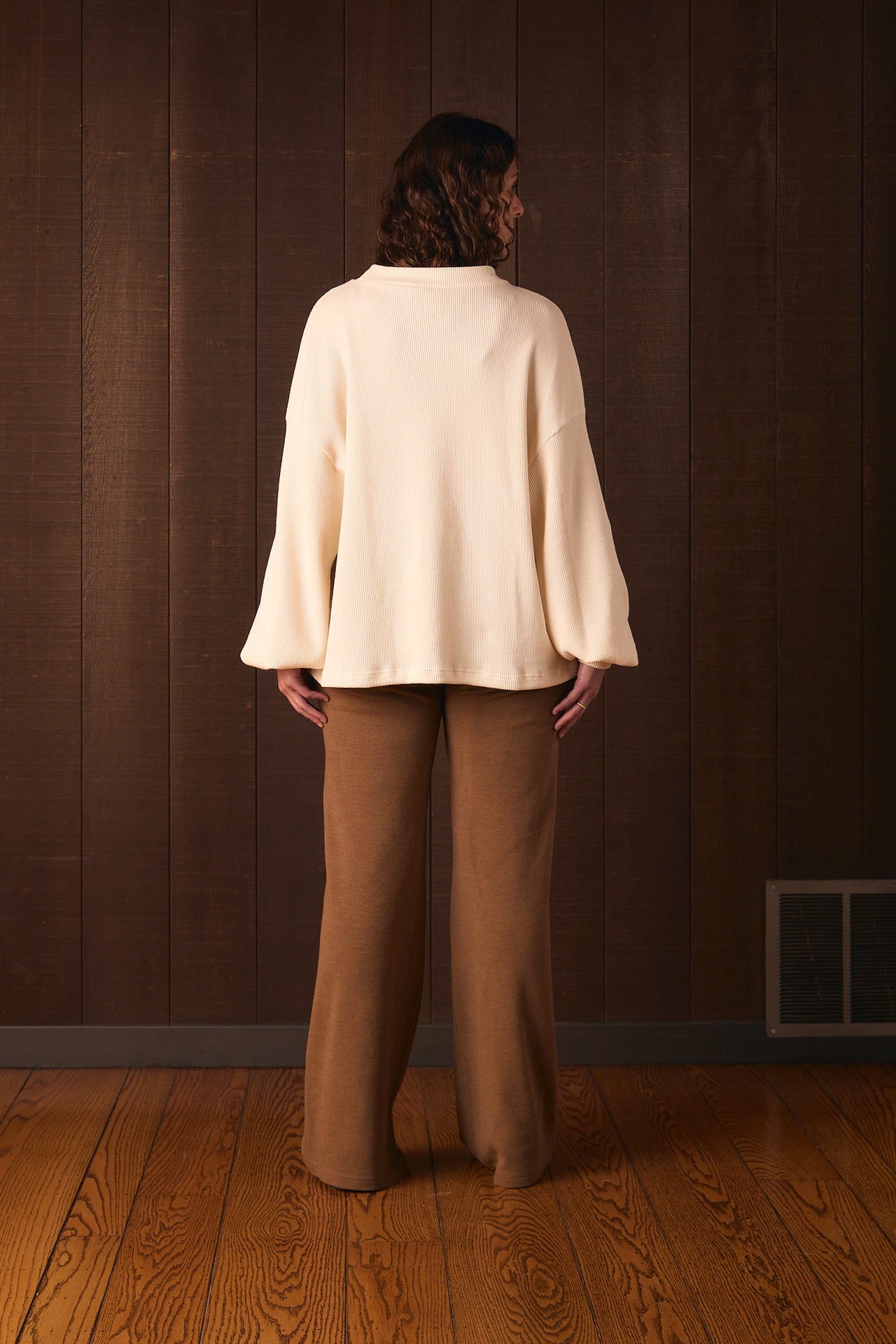 Tall woman wearing a cream sweater and brown pants standing against a wooden paneled wall.