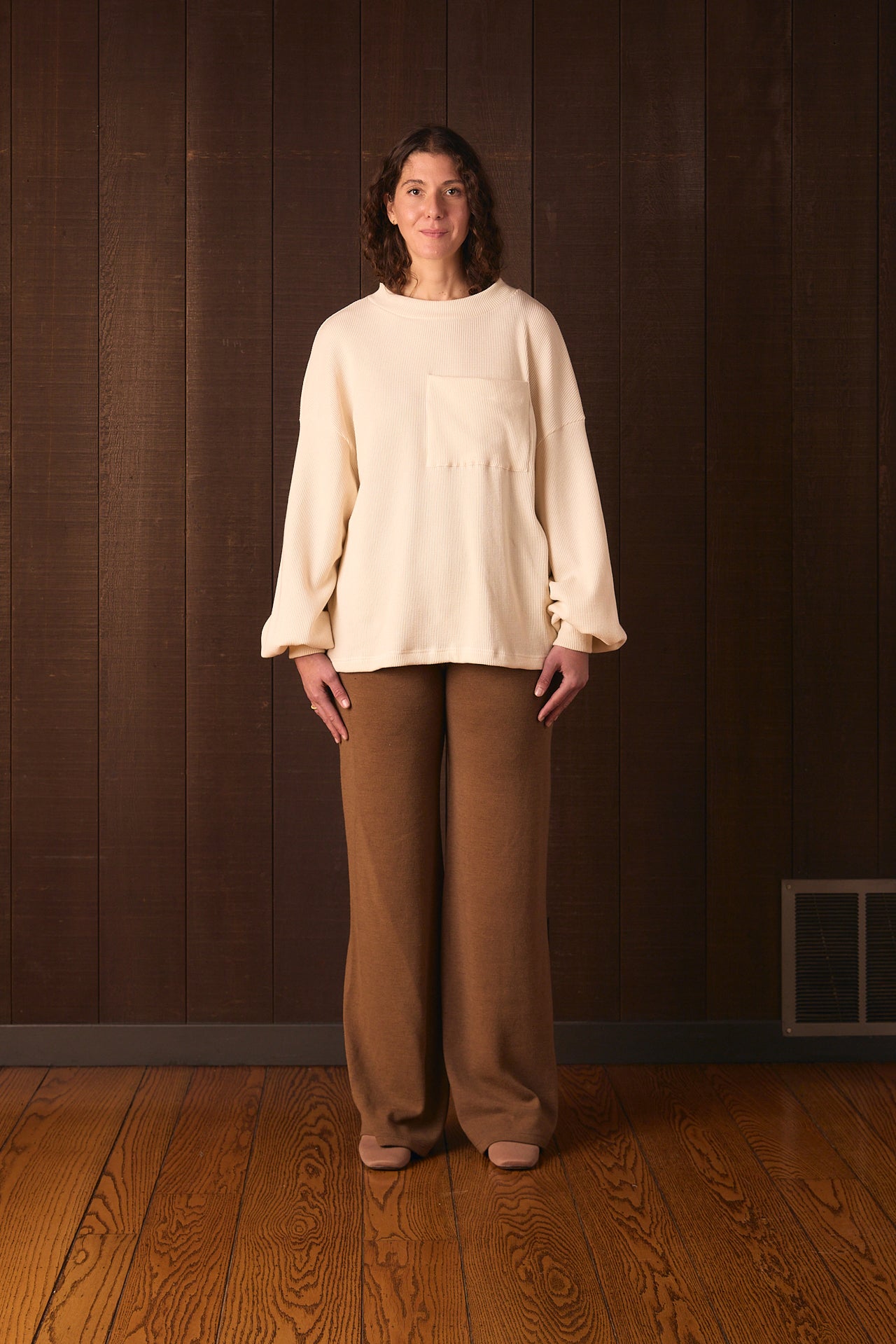 Tall woman wearing a cream sweater and brown pants standing against a wooden paneled wall.