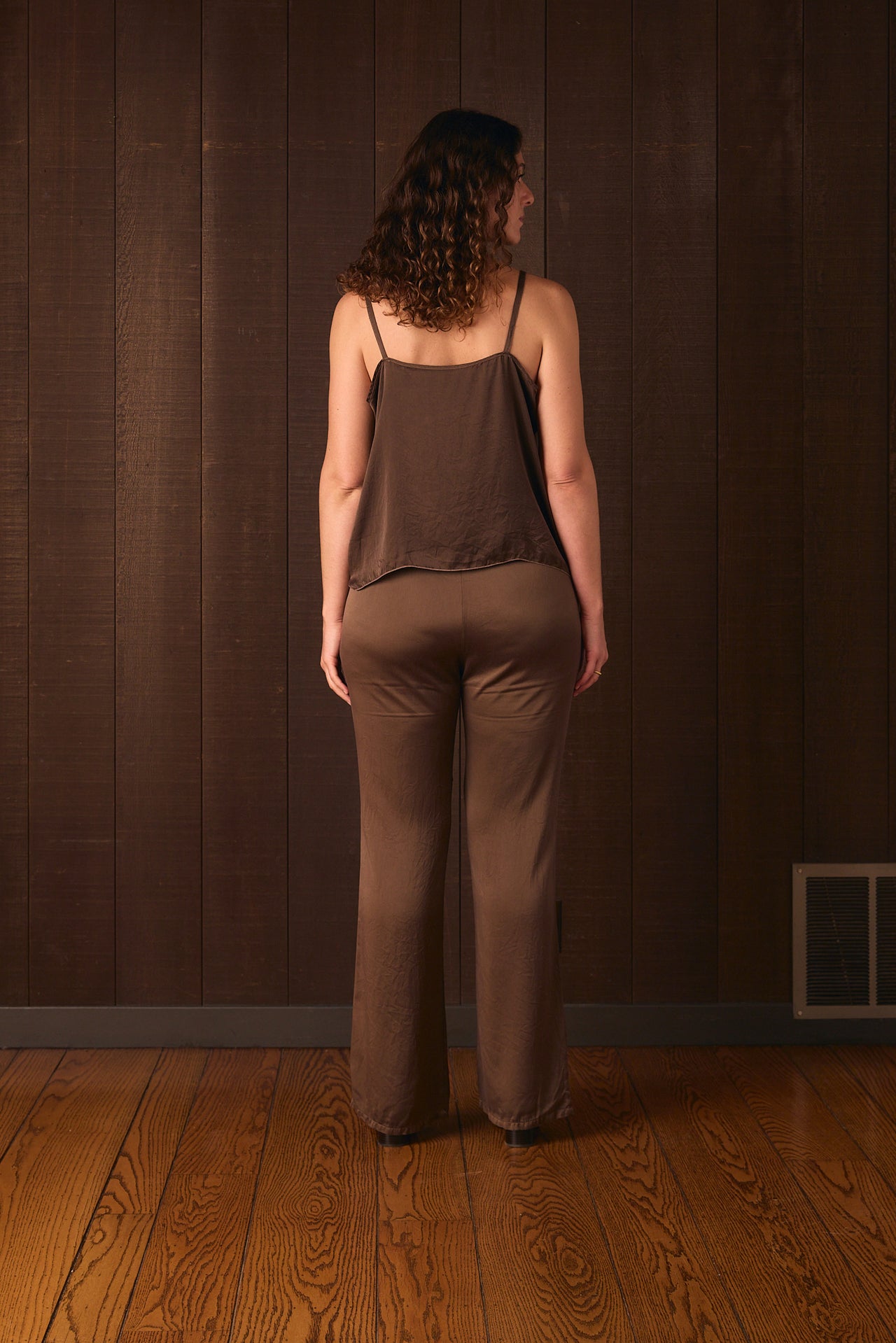 Tall woman wearing a brown jumpsuit standing against a dark brown wall.