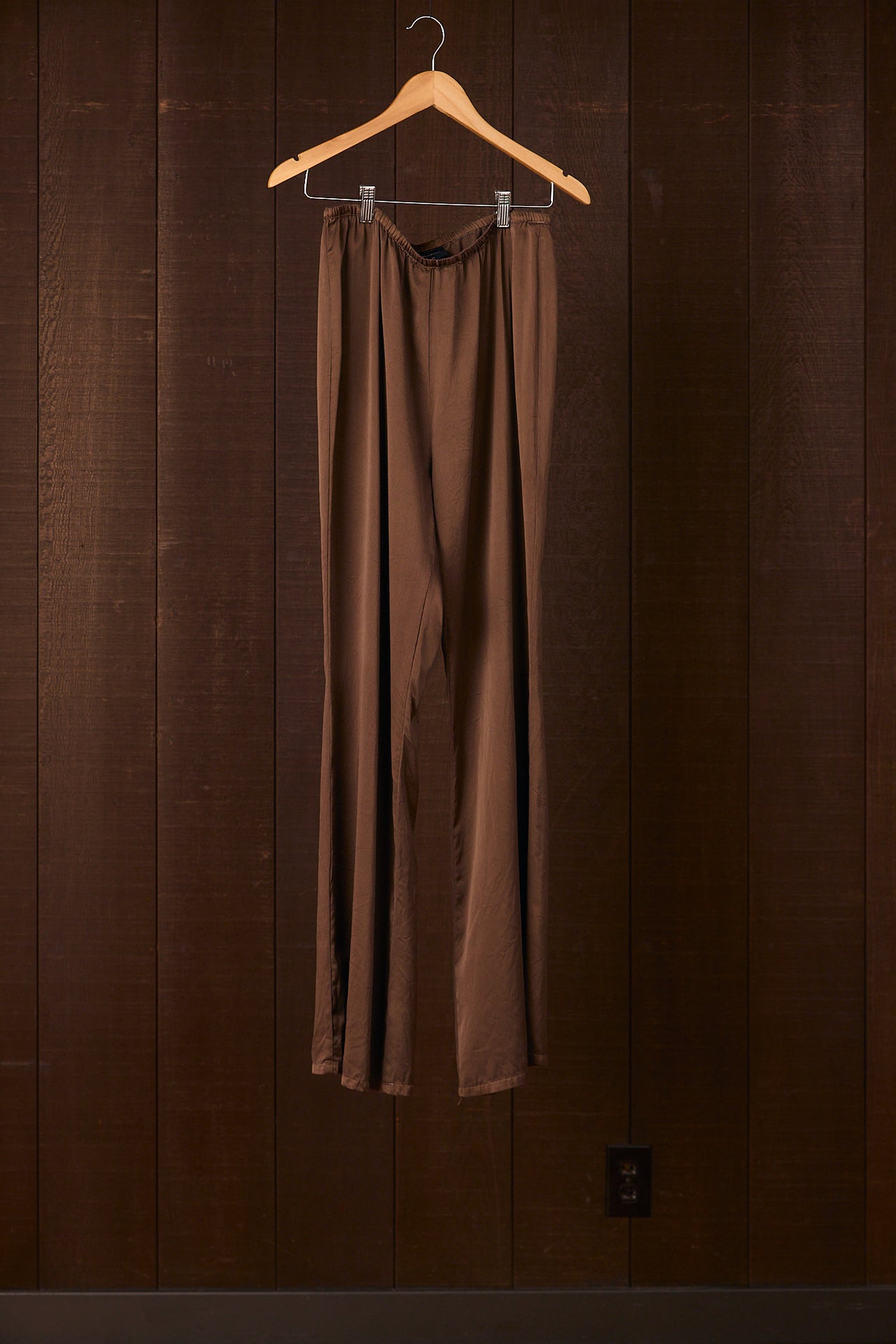 Brown silk pants hanging on a wooden hanger against a brown wall.