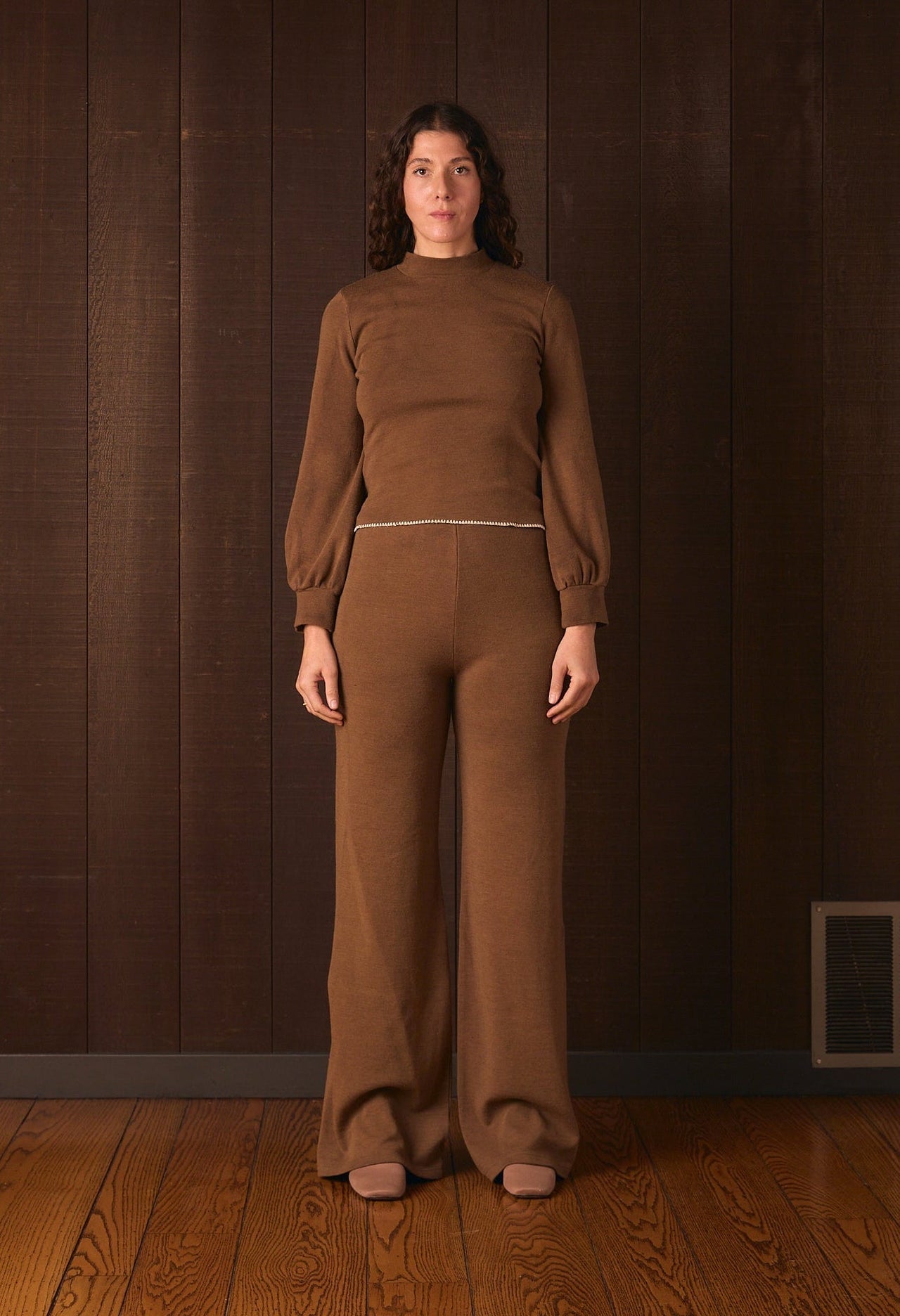 Tall woman wearing a brown outfit standing against a dark brown wall.