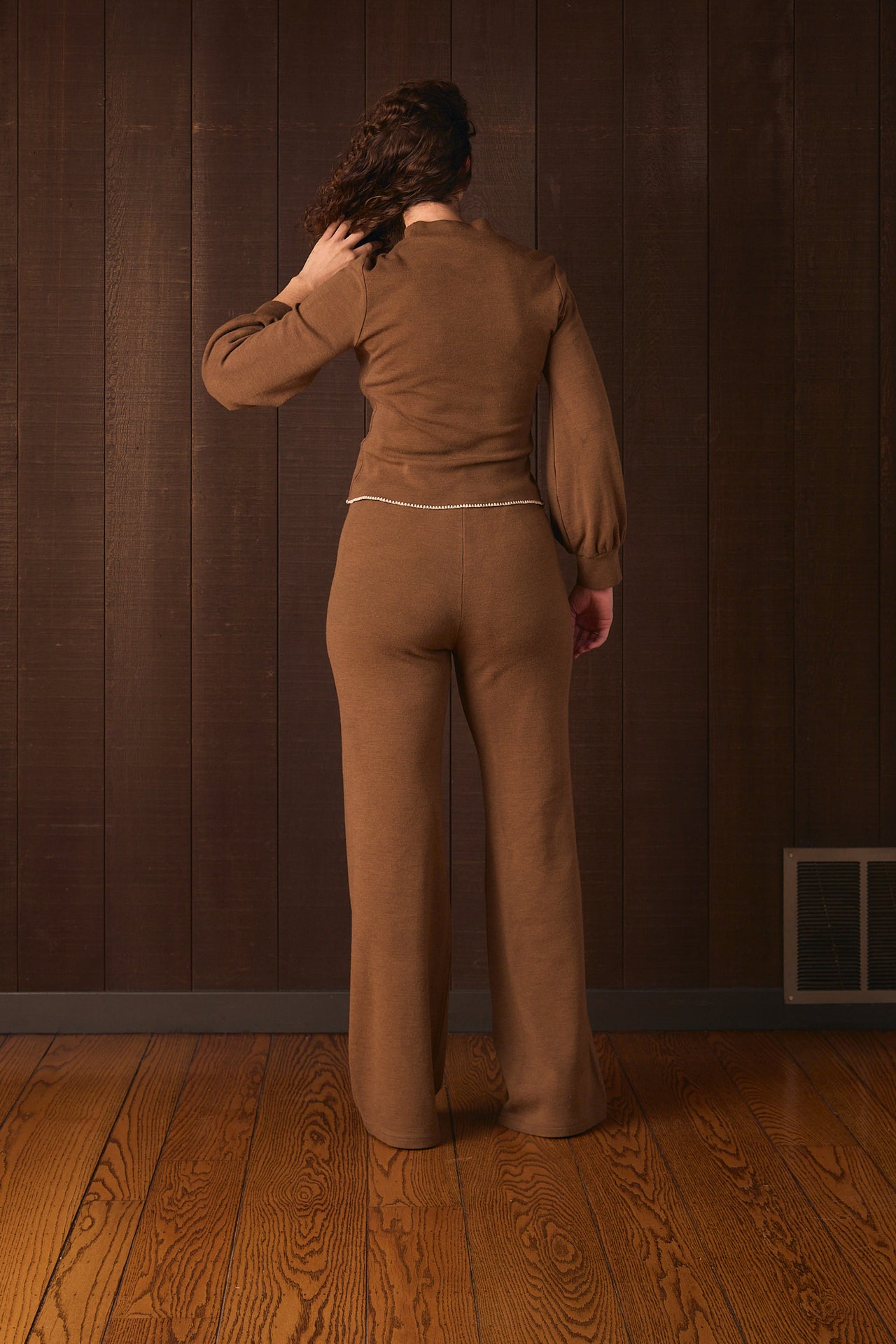 Tall woman wearing a brown outfit standing against a wooden paneled wall.