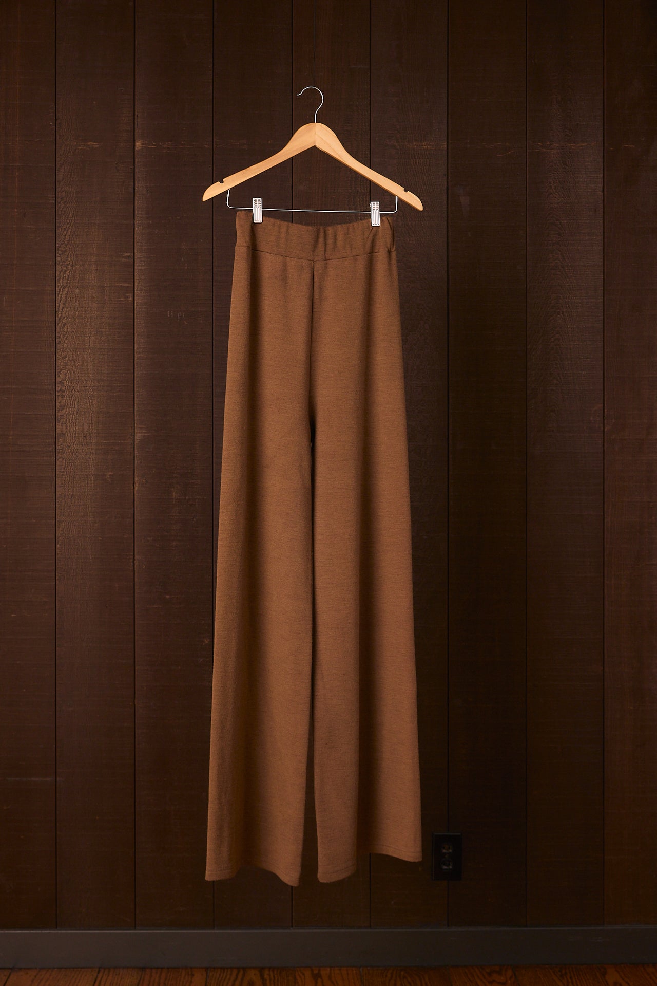 Tall women's brown pants hanging on a hanger against a dark brown wall.
