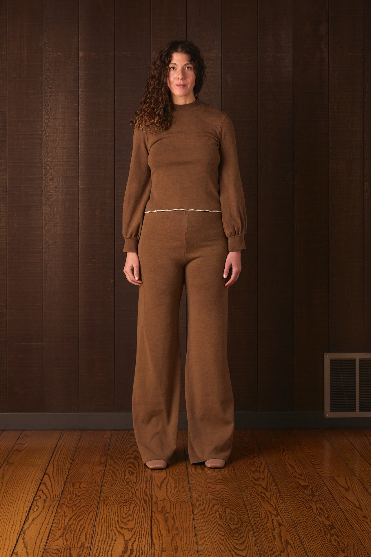 Tall woman wearing a brown outfit standing against a wooden paneled wall.