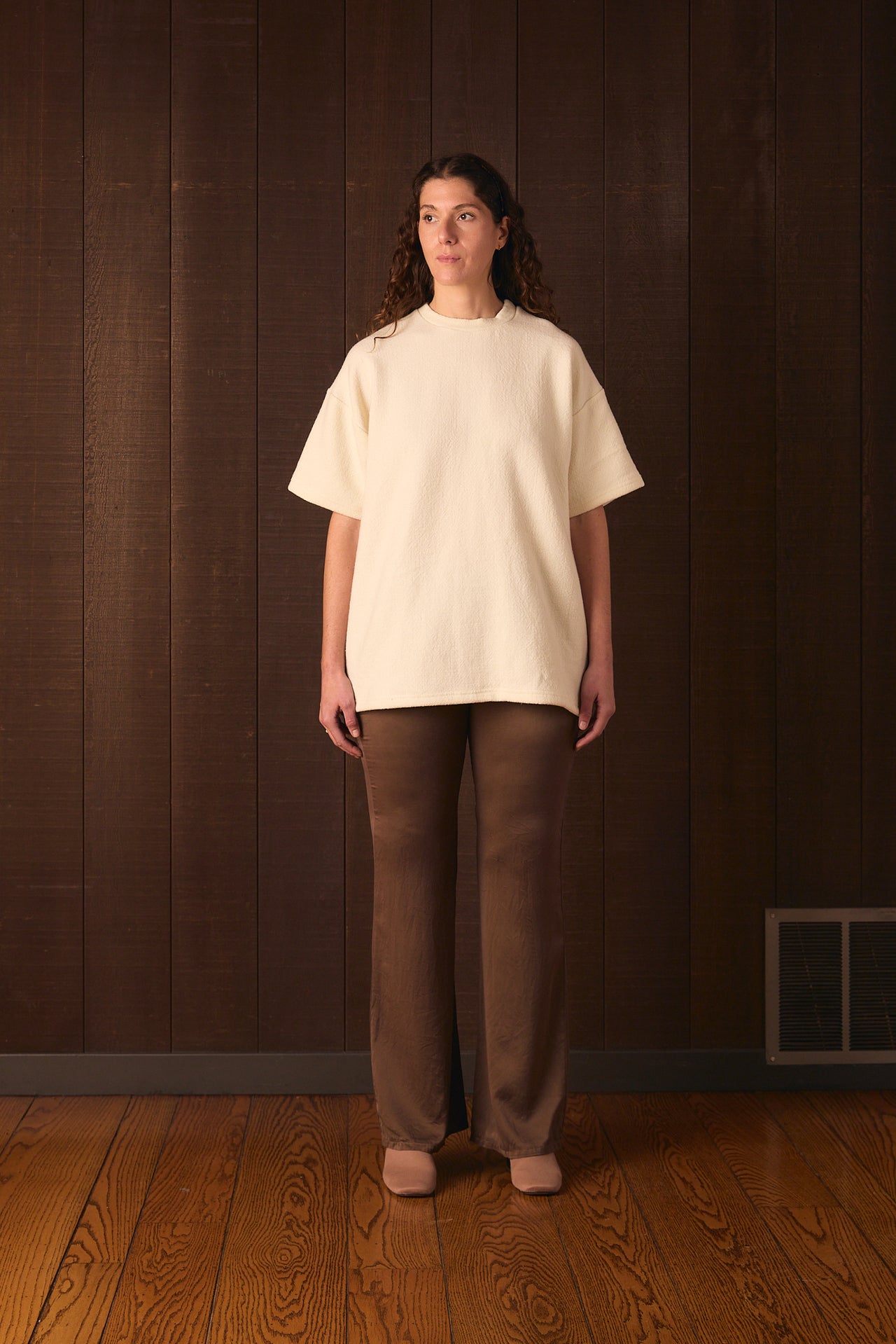 Tall Woman wearing a cream sweater and brown pants standing against a wooden paneled wall.