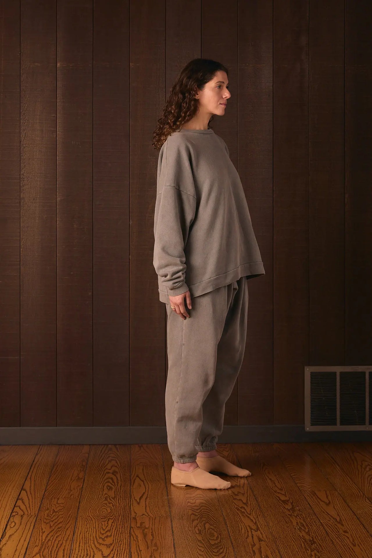 Tall woman wearing a gray outfit standing in a room with wooden flooring and a brown wall.