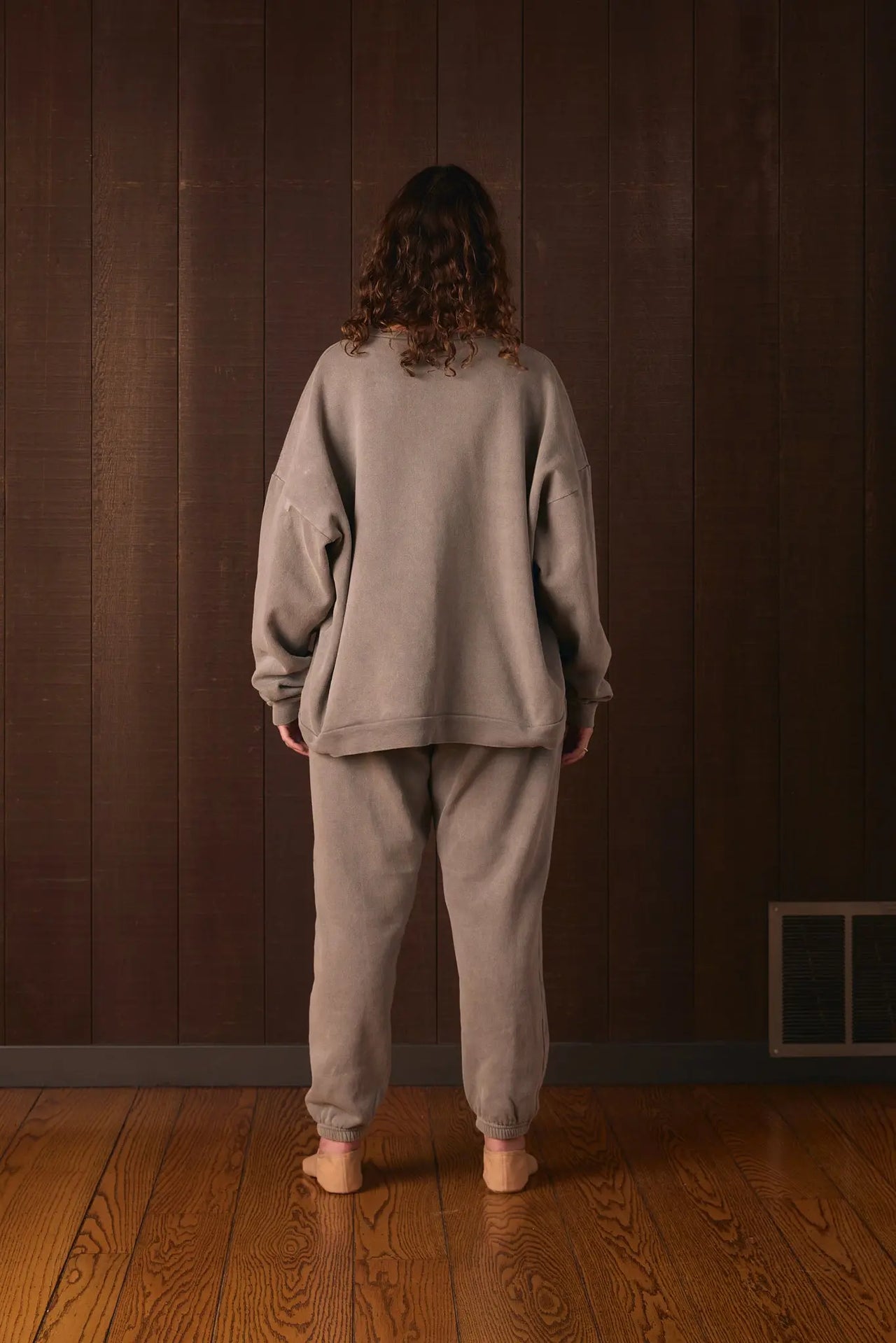 Tall woman wearing a gray tracksuit standing against a brown wall.