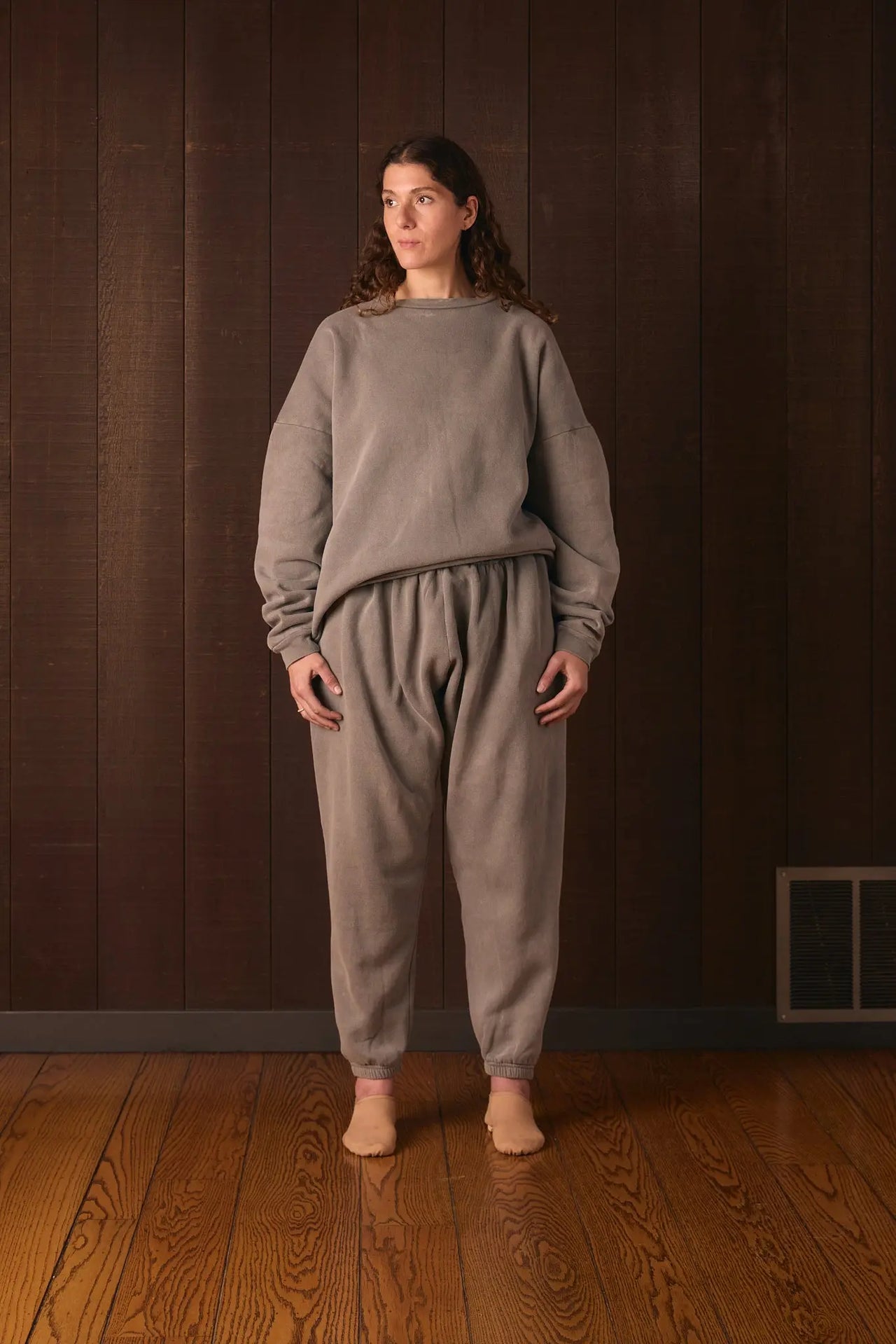 Tall woman wearing a gray tracksuit standing against a dark brown wall.