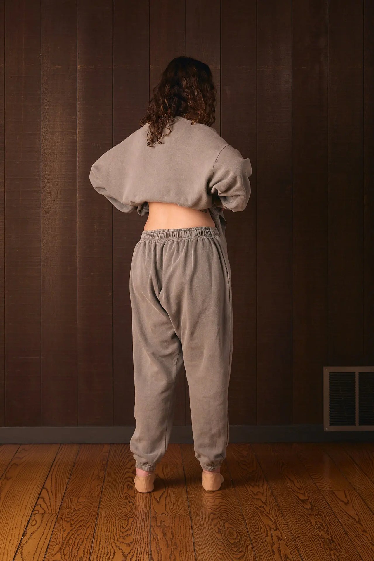 Tall woman wearing a gray sweatshirt and pants standing against a brown wall.