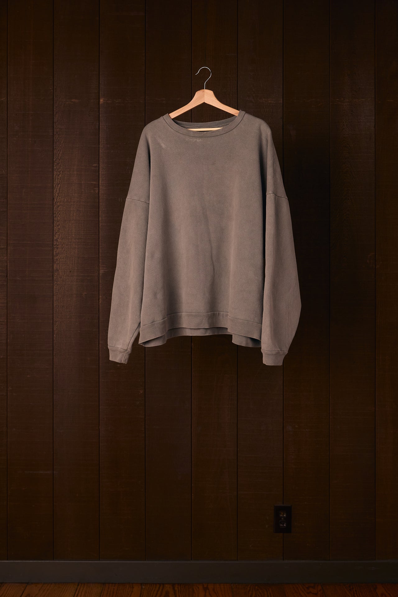 Tall women's gray sweatshirt hanging on a wooden hanger against a dark wooden wall.