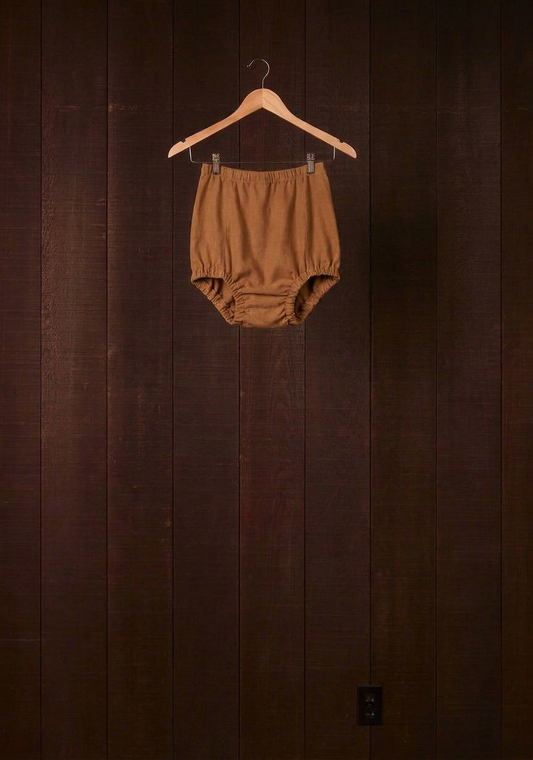 Cutch plant-dyed walnut cotton Comfort Bloomer with scrunchie leg finish on a wooden hanger against a dark wood wall
